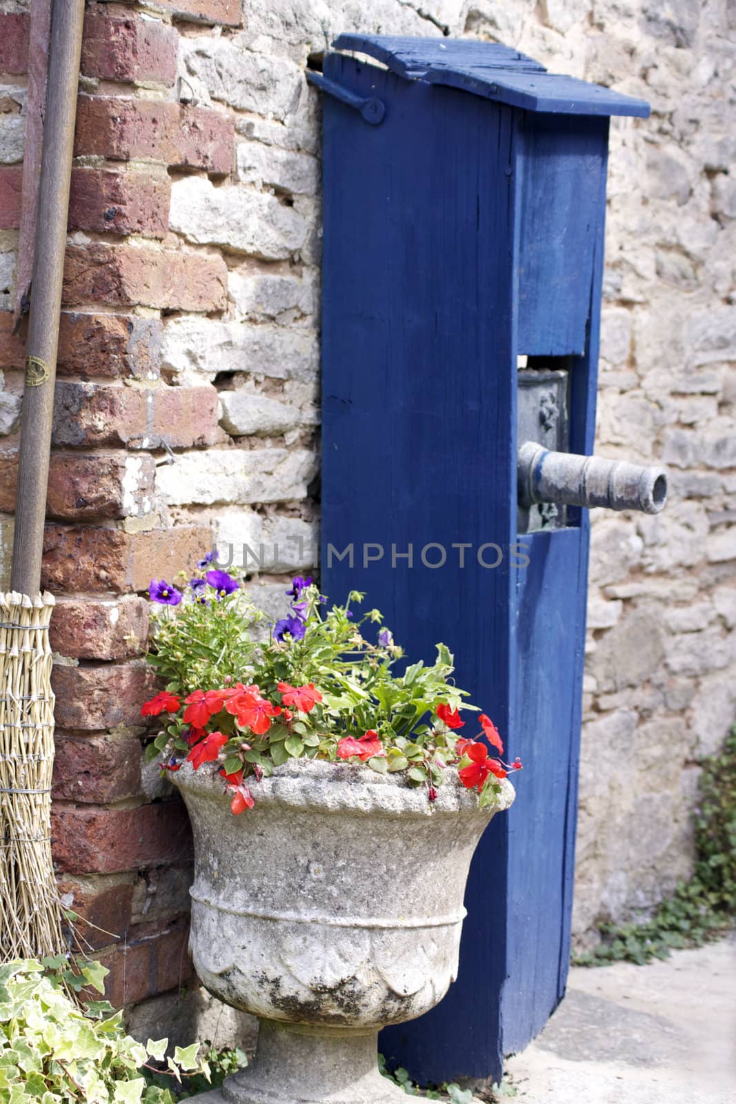 An old water pump covered in a blue painted wooden constructed cover, next to a stone urn containing summer pansies. Set against an old brick wall.