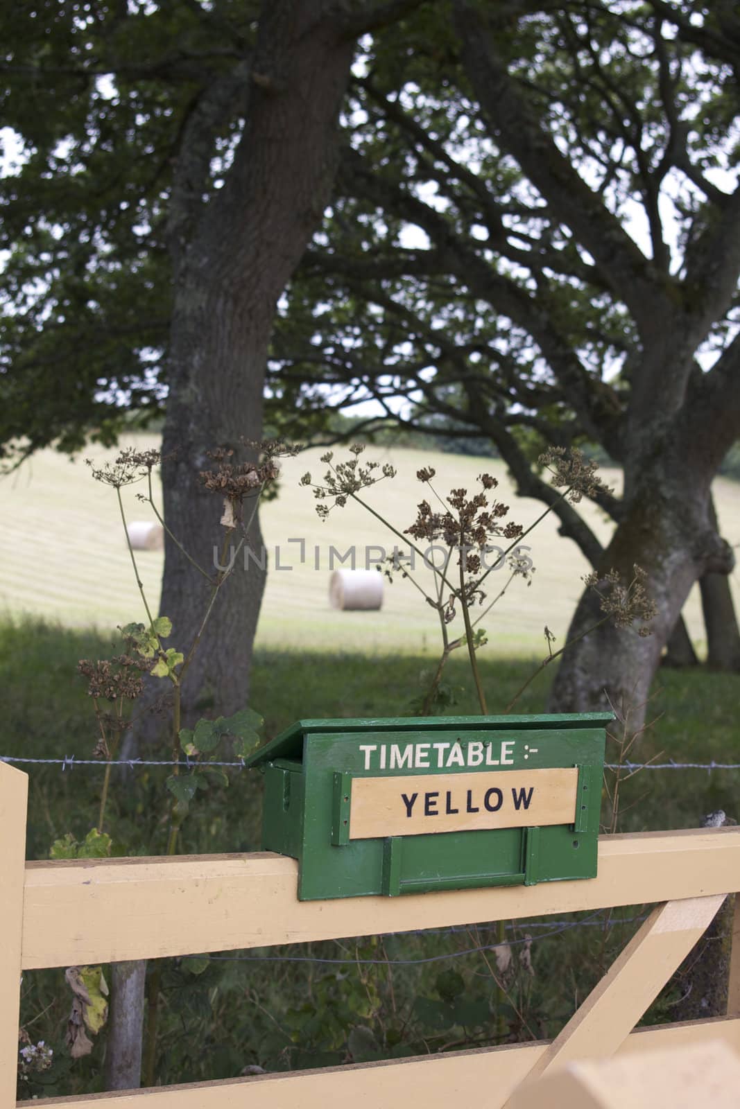 A timetable sign constructed of wood and painted green, set on top of a cream painted fence. Rolling fields with straw bails set to the background. This forms part of the notification system for which timetable is used on the Swanage Steam Railway in Dorset.