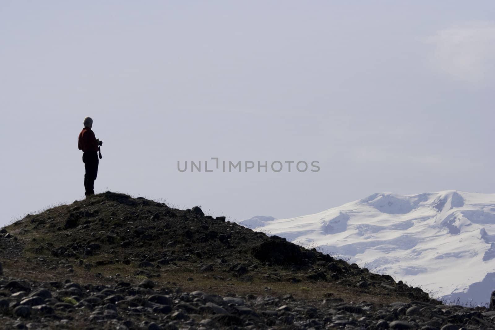 Lone traveler stands silhouetted against the sky with stony mountain beneath his feet and snow and ice in his view