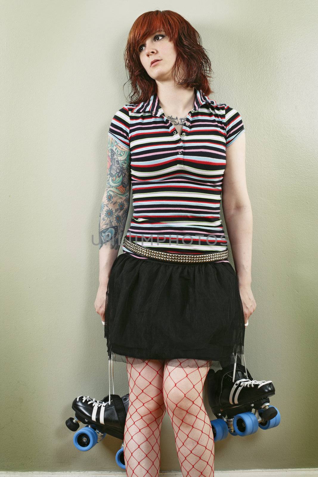 Photograph of a bored roller derby girl holding her skates and waiting against a wall.