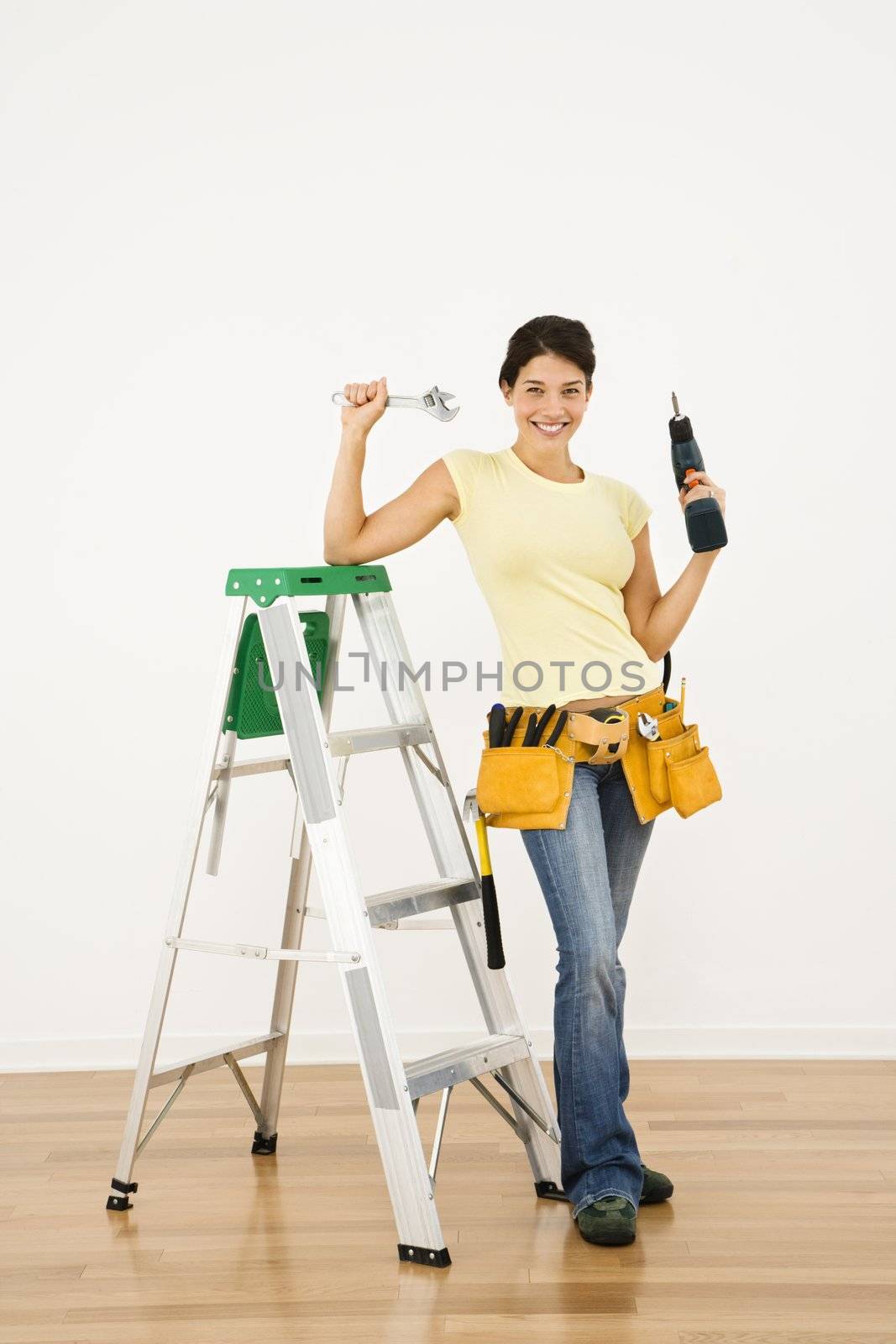 Woman standing in home with ladder and holding tools smiling.