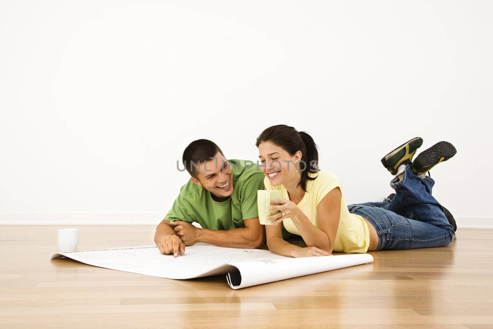 Attractive young adult couple lying on home floor with coffee cups smiling and looking at blueprints.