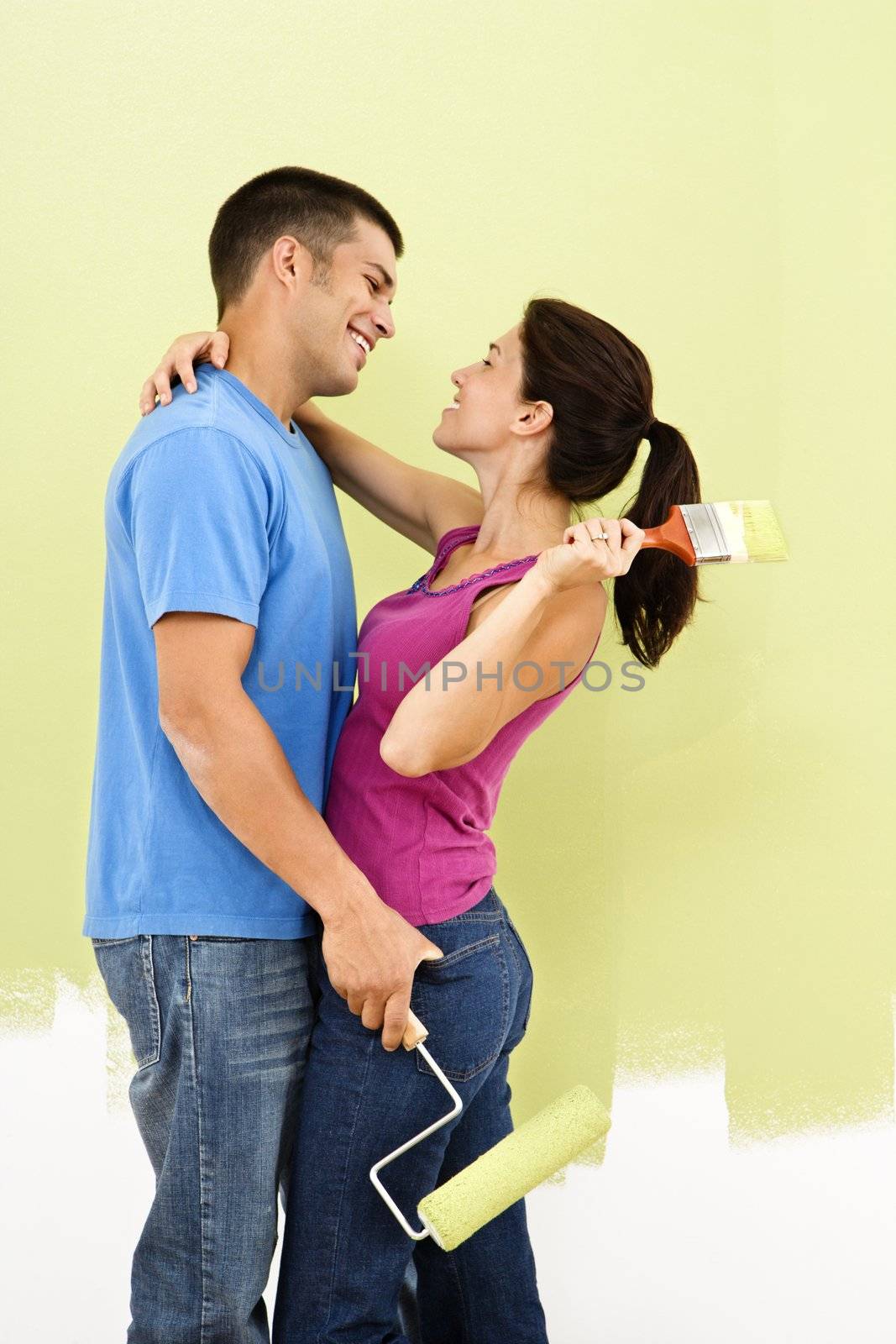 Couple hugging and smiling at eachother holding paintbrushes in front of partially painted wall.