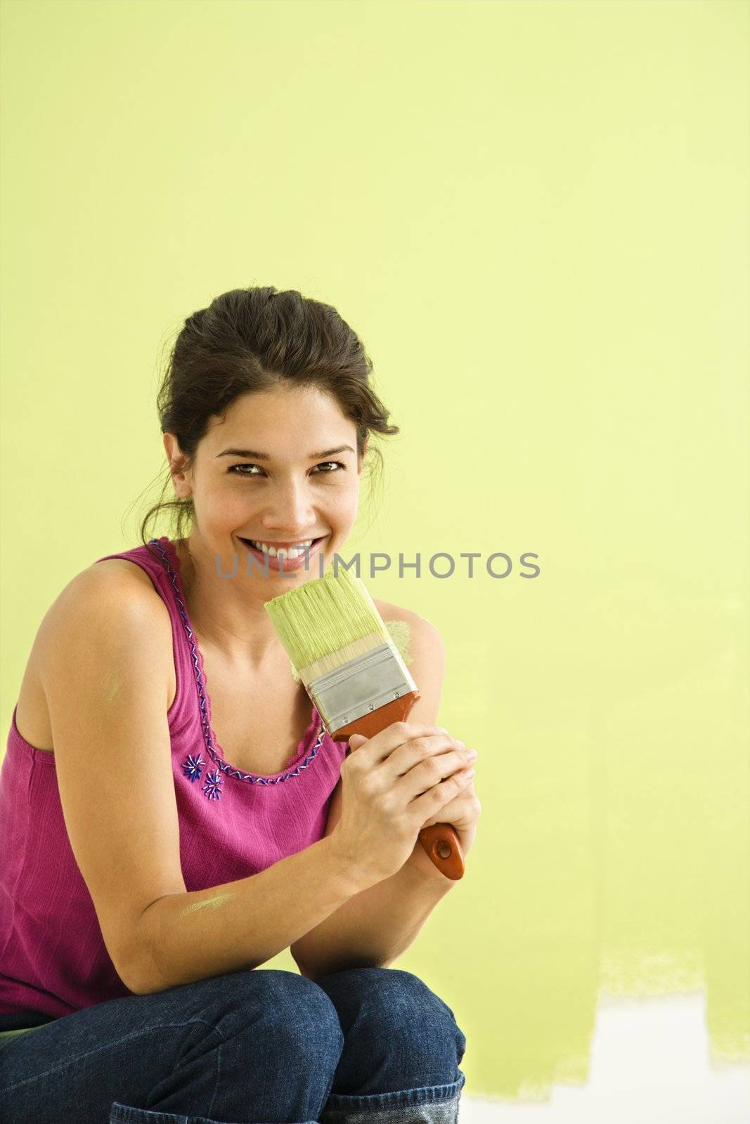 Pretty smiling woman kneeling in front of partially painted wall holding paintbrush.