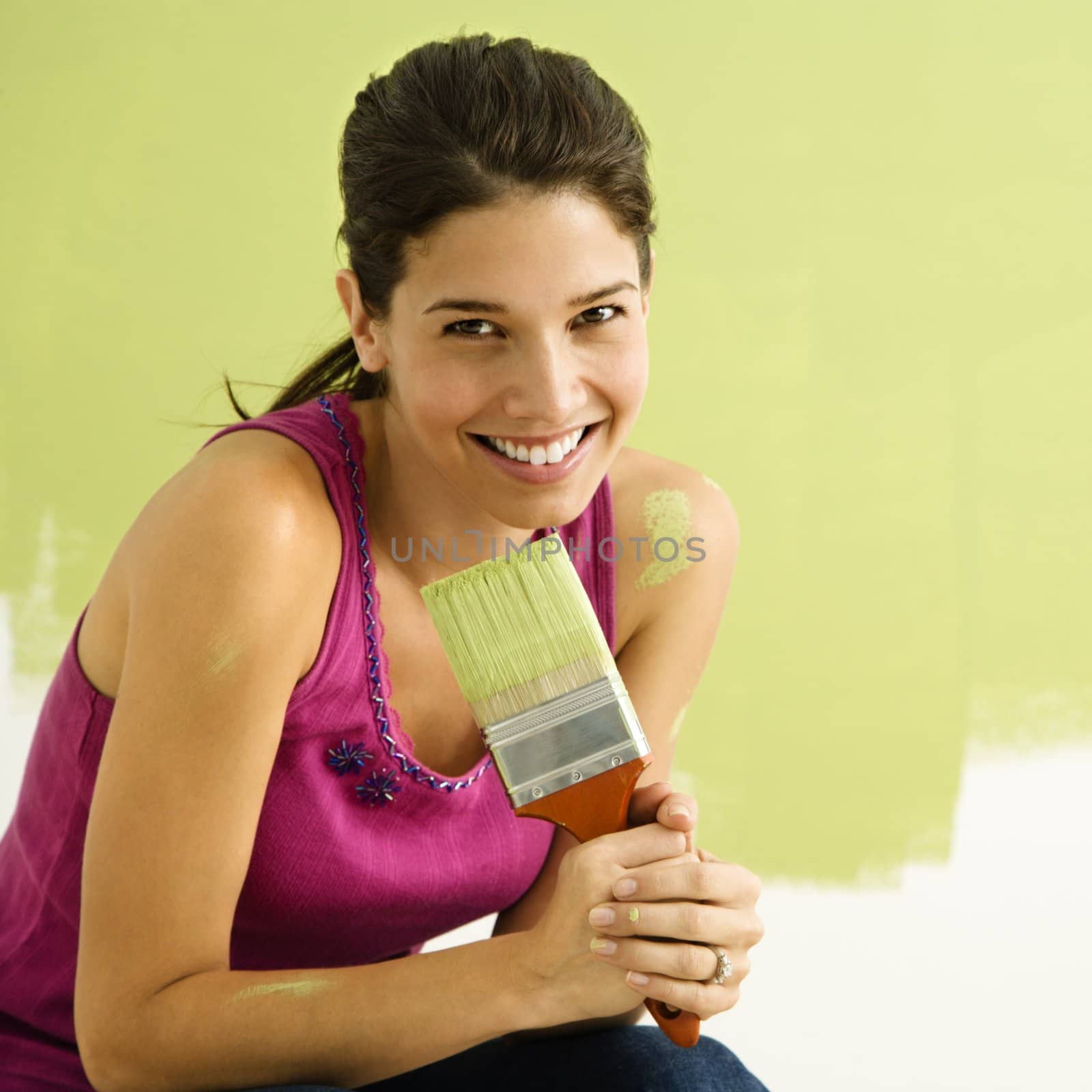 Pretty smiling woman kneeling in front of partially painted wall holding paintbrush.
