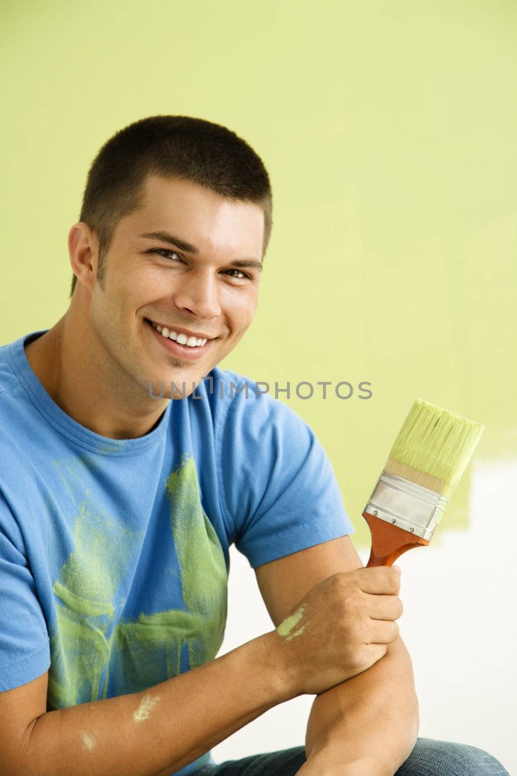 Smiling man kneeling in front of partially painted wall holding paintbrush.