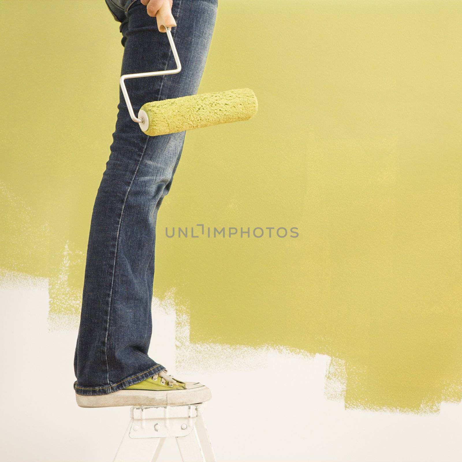 Legs of woman standing on stepladder holding paint roller with painted wall.