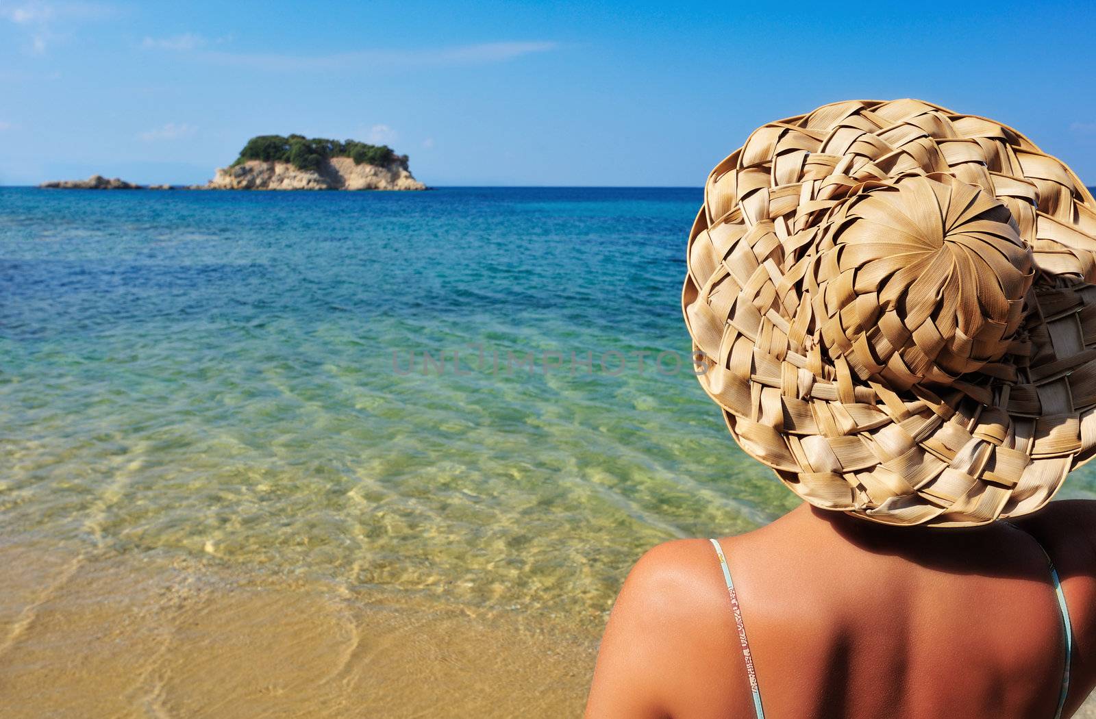 Closeup image of a young woman in a straw hat and bikini, looking at a tiny island in the Mediterranean
