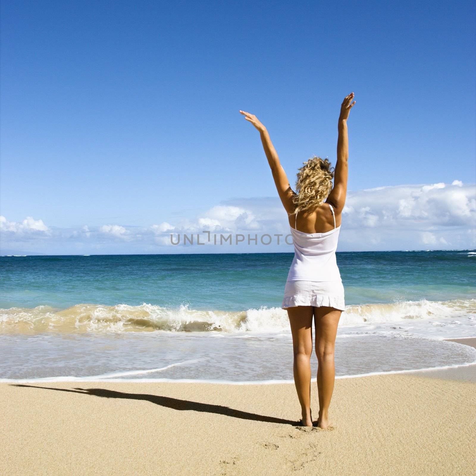 Pretty young woman standing on Maui, Hawaii beach with arms raised into air looking out towards ocean.