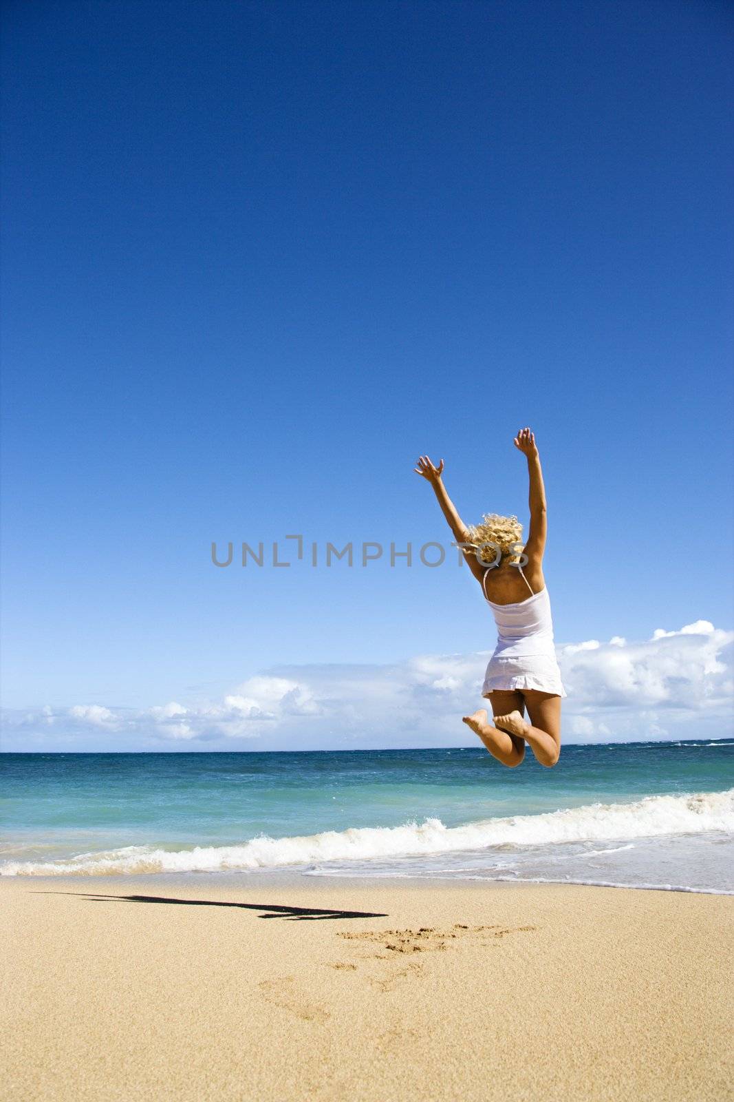 Pretty young woman jumping on Maui, Hawaii beach with arms raised into air.