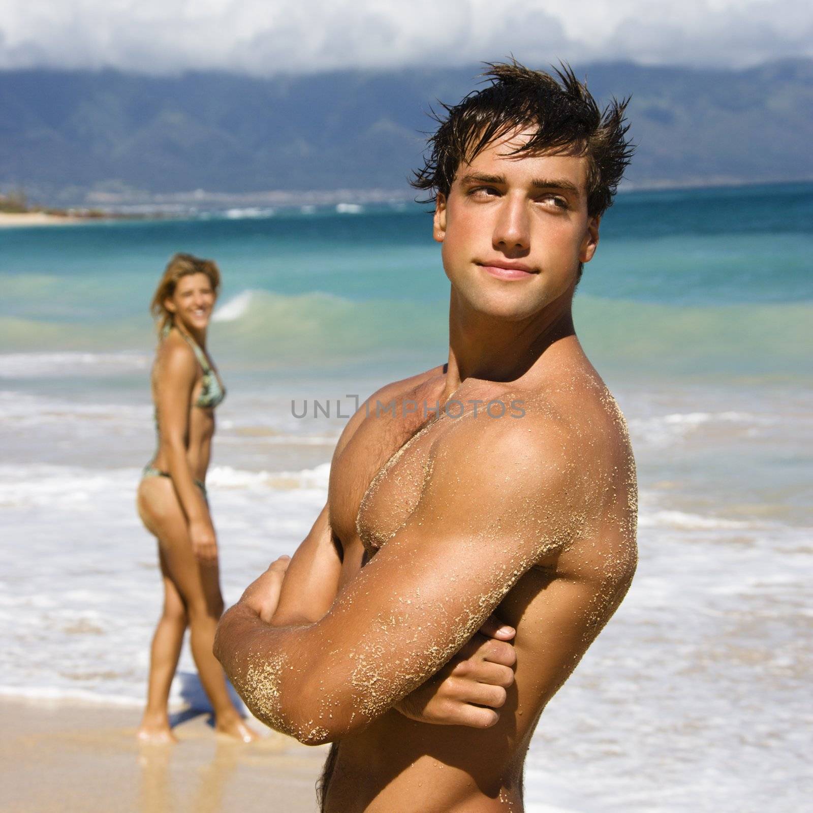 Handsome man standing on Maui, Hawaii beach with woman in background.