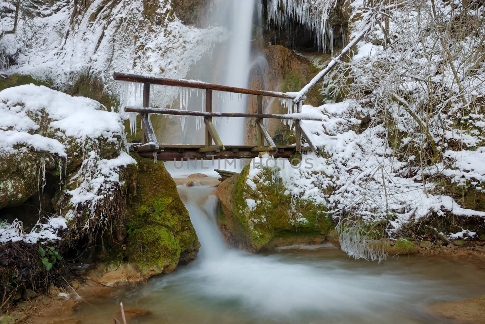 Waterfall with icicles and snow around with a small wooden bridge on a winter day.