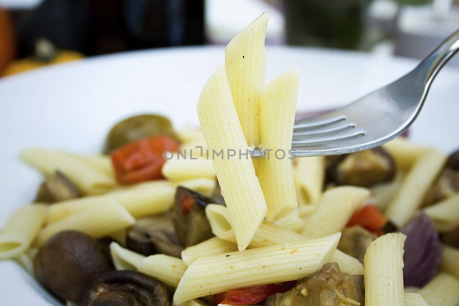 Several pieces of penne pasta on fork with gourmet pasta dish in background.  