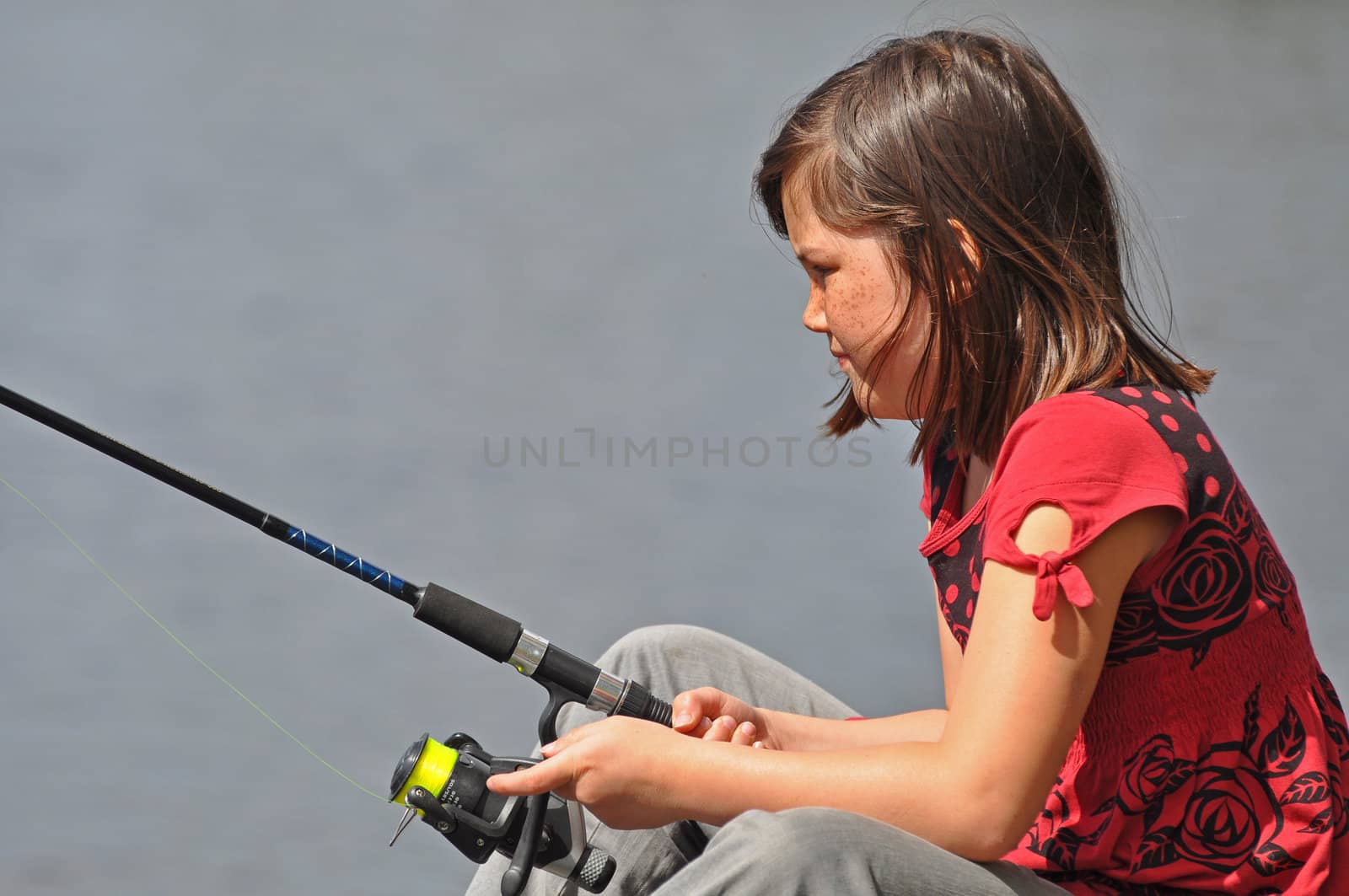 A young girl sitting by a lake with a fishing rod in her hands.
