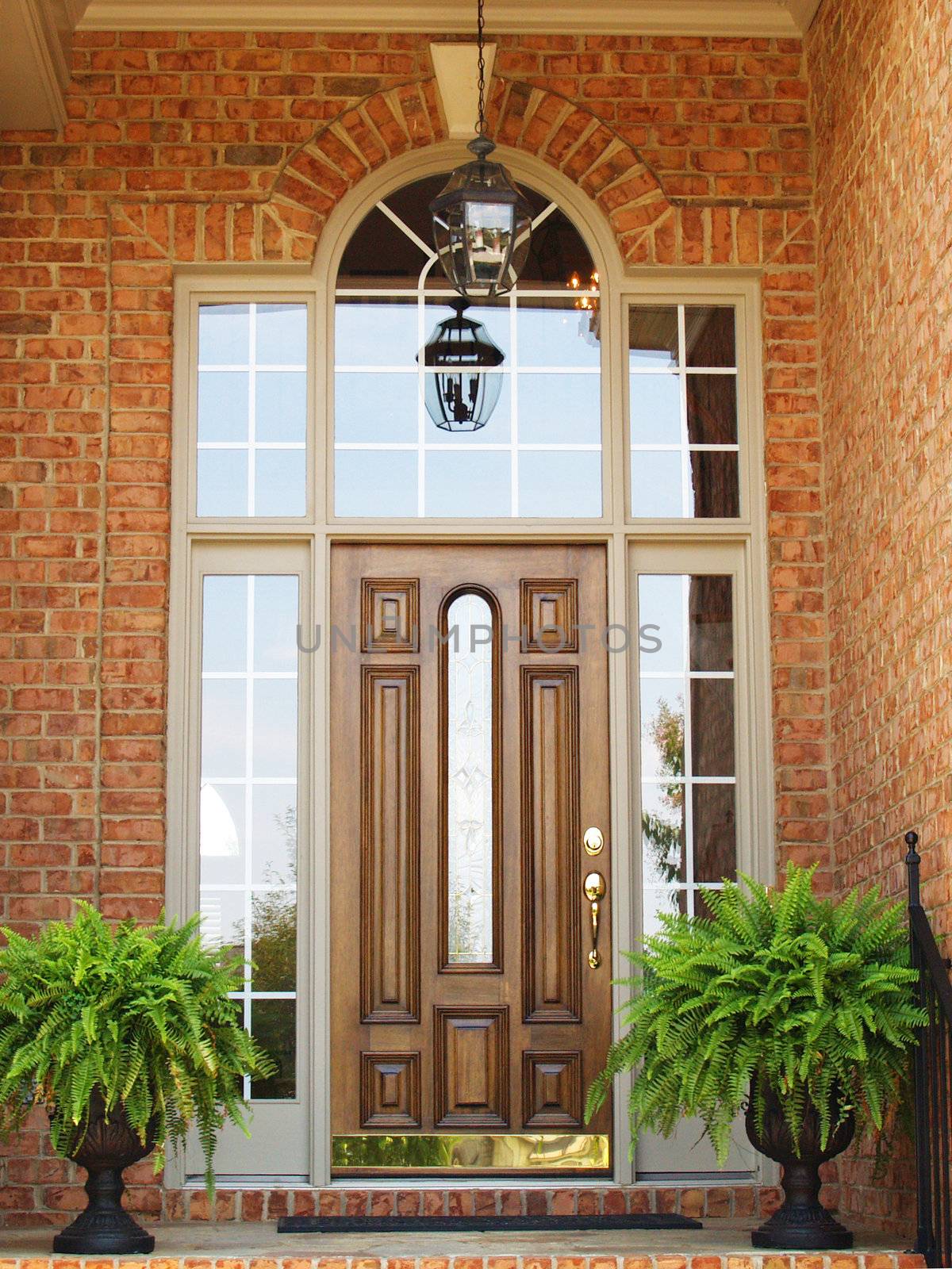 Front door of a brick luxury home with a wooden door, hanging light and planters with ferns.