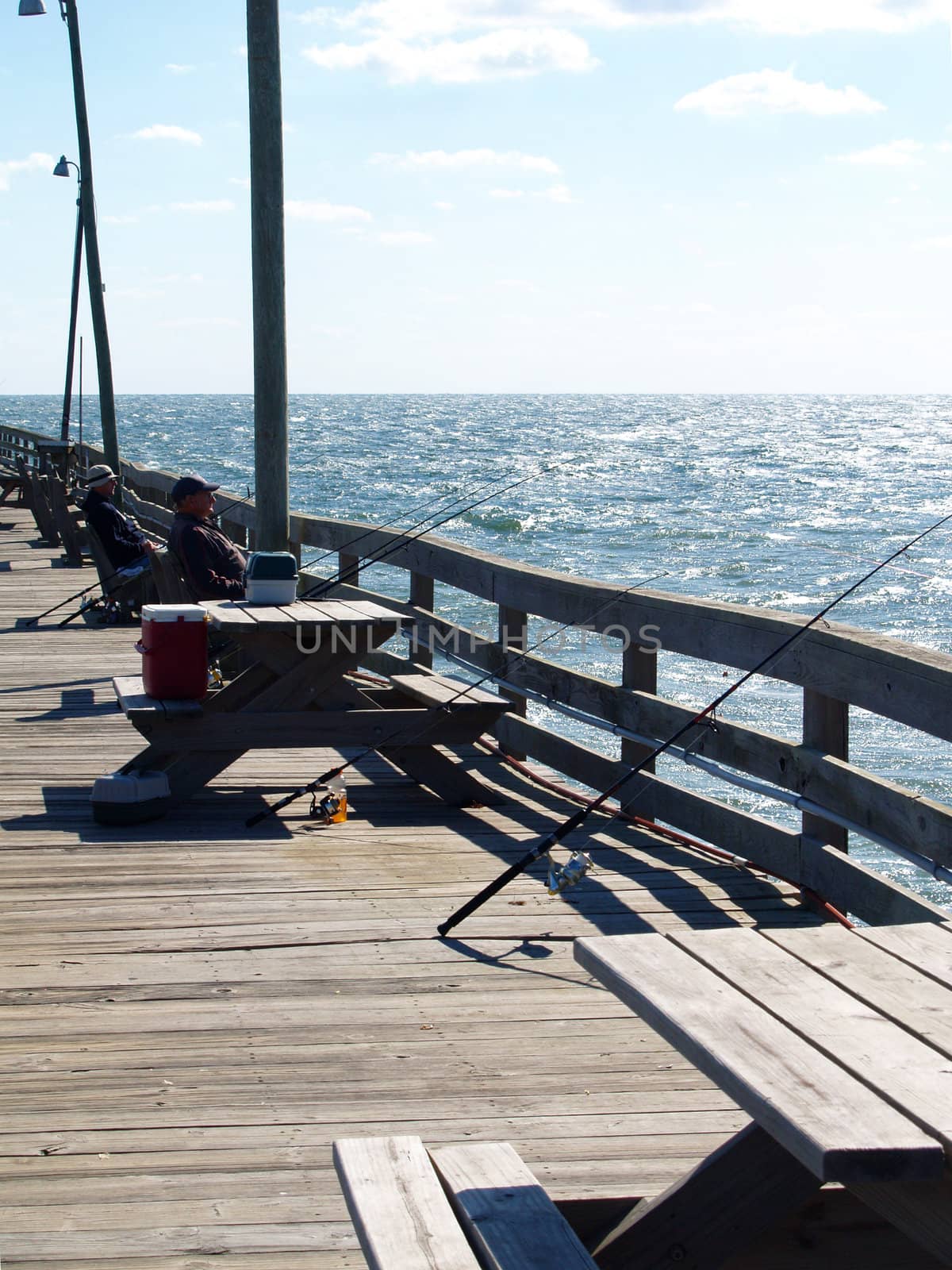 Two men fishing from a pier over the Atlantic Ocean. They have fishing rods, coolers, and other assorted equipment.