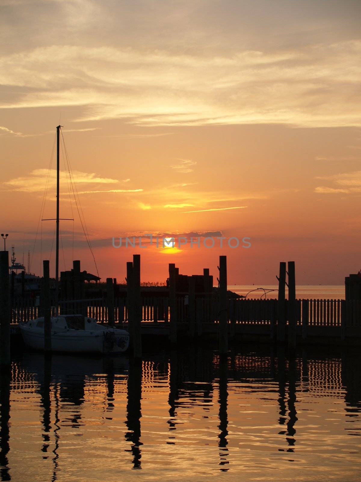 Summer sunset over a maritime marina. A lone sailboat docked casts a reflection in the rippling water.