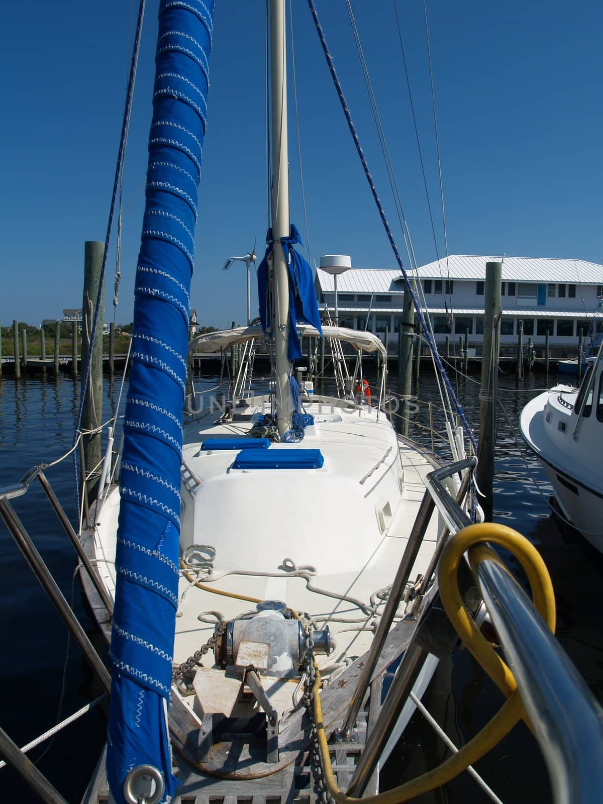 Abstract view from the bow of a sailboat at the dock with the rigging, railings and marina pilings visible. A bright blue sail wrapped around the mainstay is in the forground.