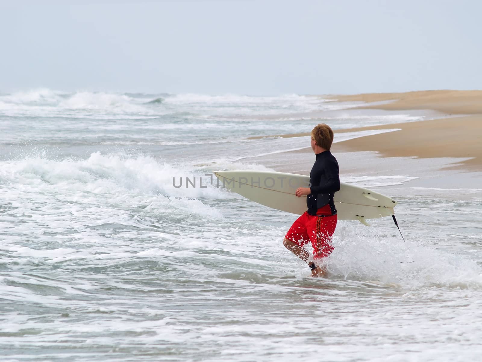 Young man with surfboard wades into the water on empty beach