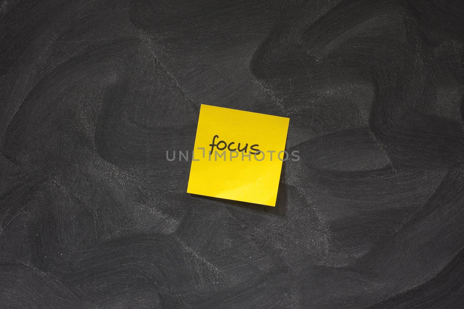 yellow sticky note with word focus posted on blackboard with chaotic white chalk eraser patterns