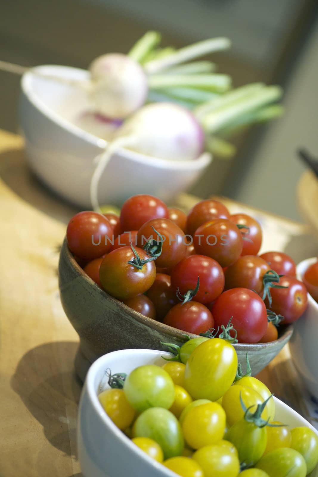 A bowl of freshly picked organic red tomatoes and a bowl of yellow pear shaped tomatoes set in a kitchen setting on top of a wooden table top. A white bowl containing freshly picked turnips in soft focus to the background.