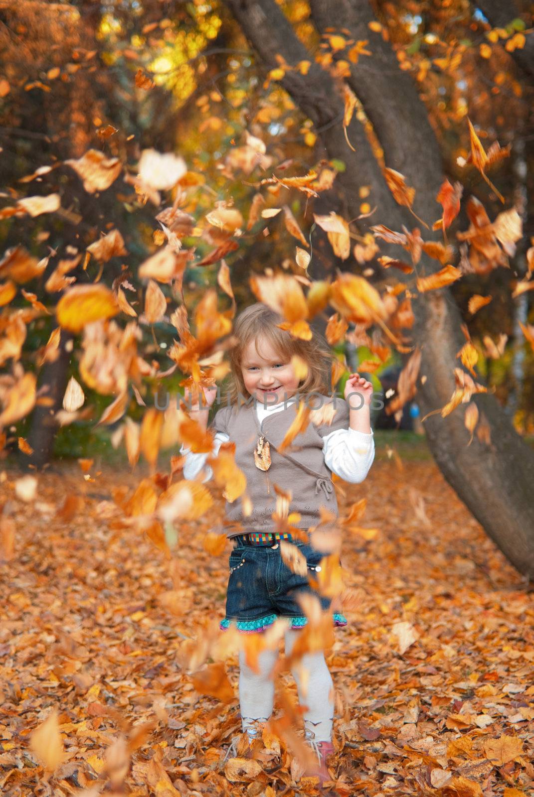 Small girl with smile (3 years old) in casual clothes is playing with yellow autumn leaves in the park.