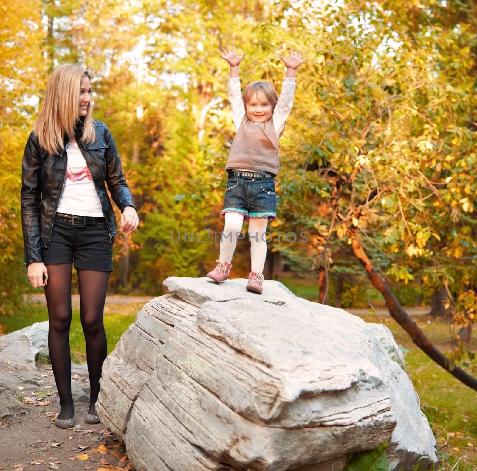Mother and daughter (3 years) are walking in autumn park in casual clothes. Girl stands on rock with smile and opened hands