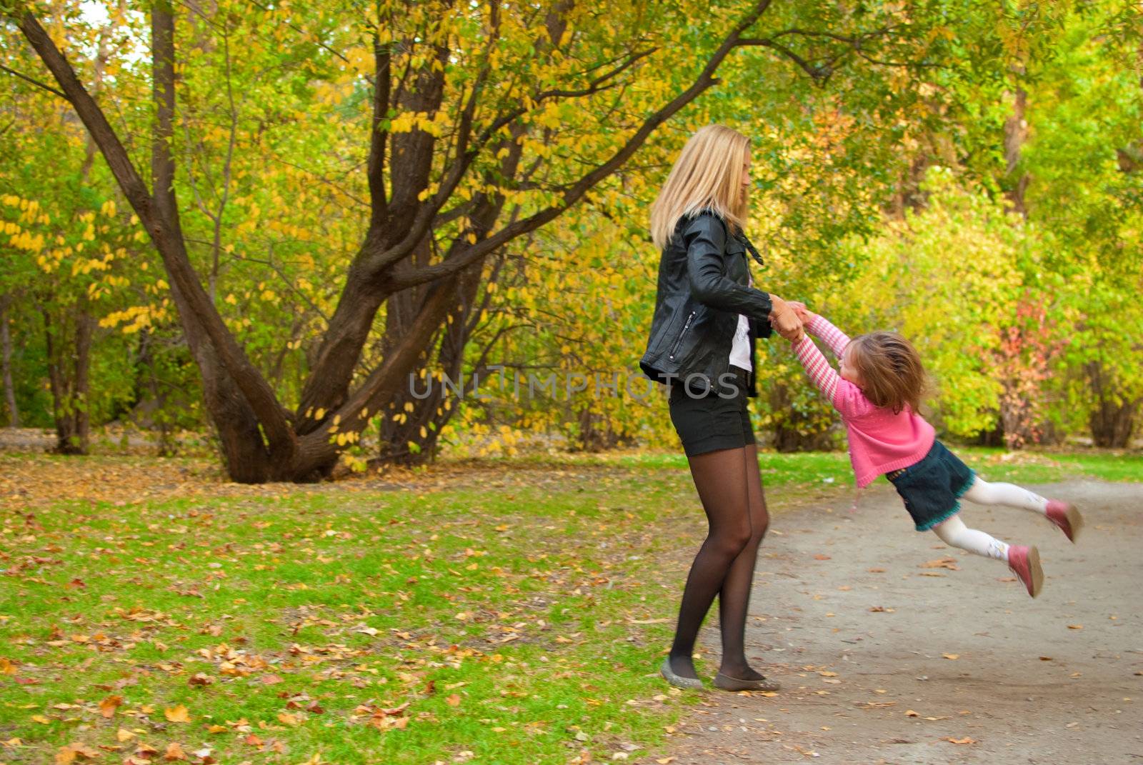 Mom is spining around with her daughter (3 years old) in autumn park.