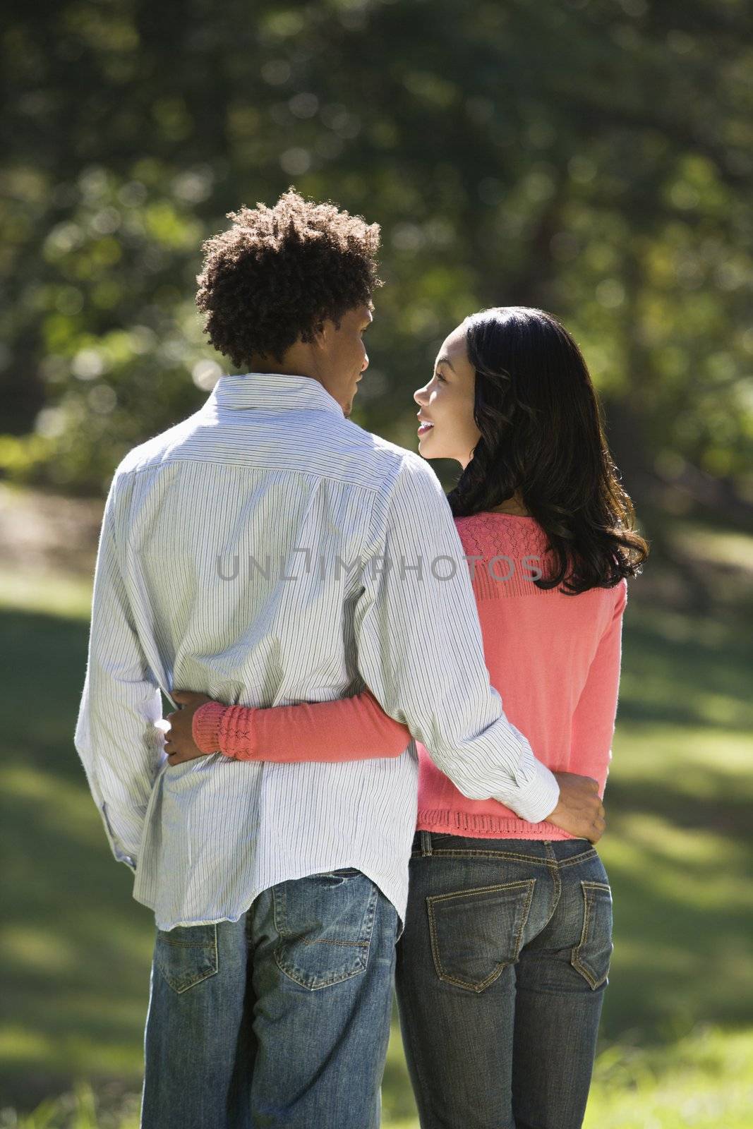 Rear view of couple with arms around eachother looking into eachother's eyes in park.