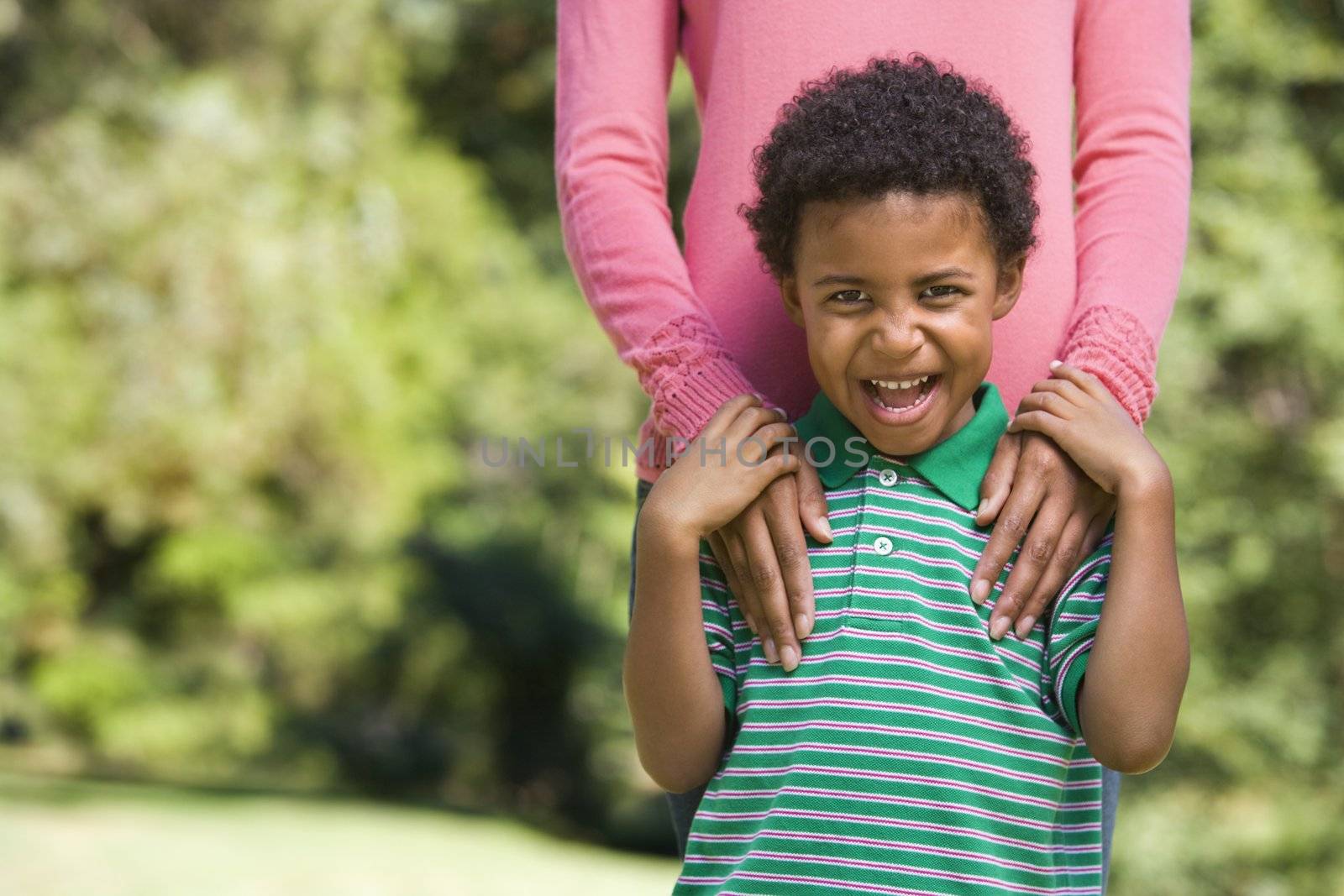Boy in park making funny expression with mother's hands on shoulders.