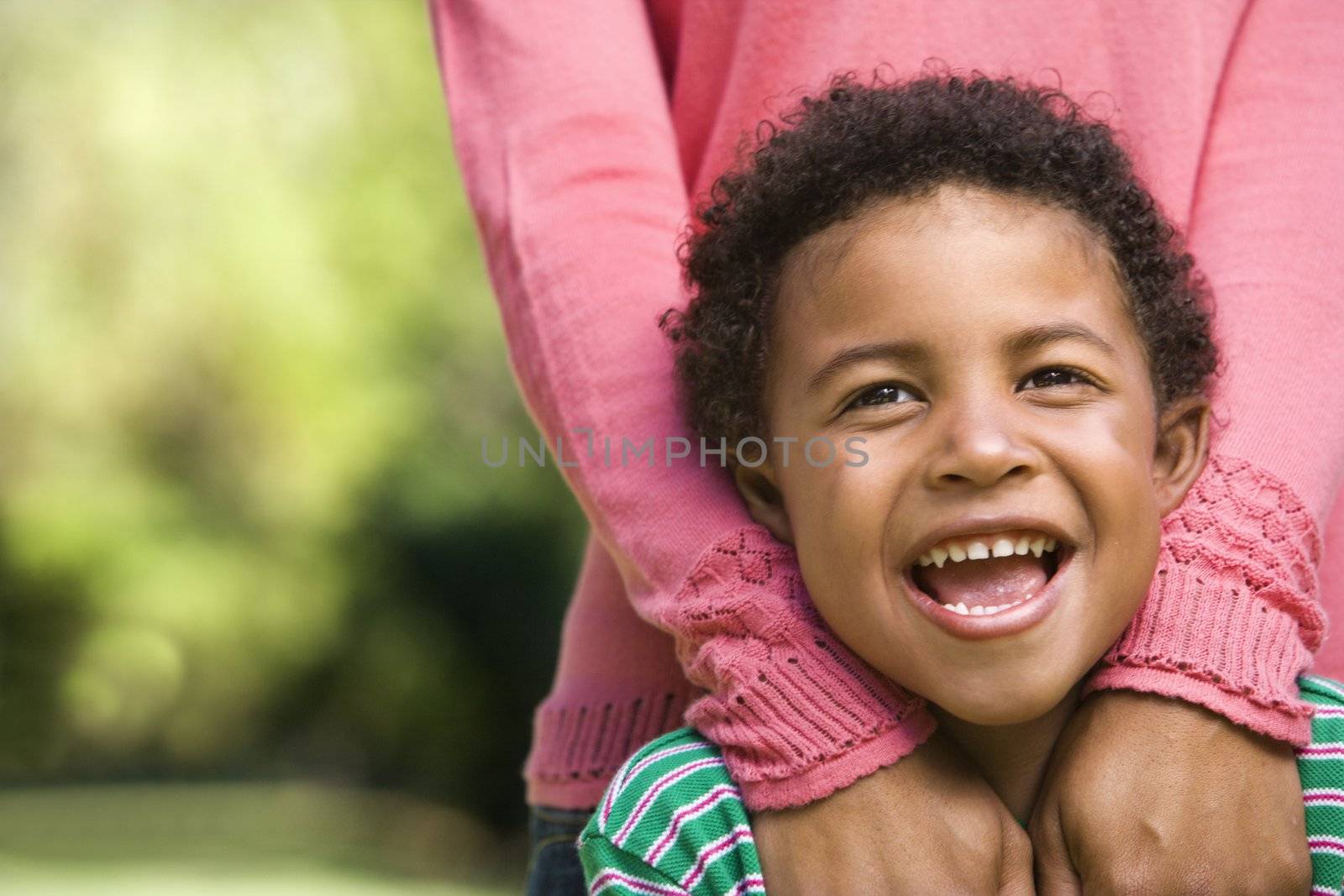 Boy smiling with mother standing behind him with hands on shoulders.