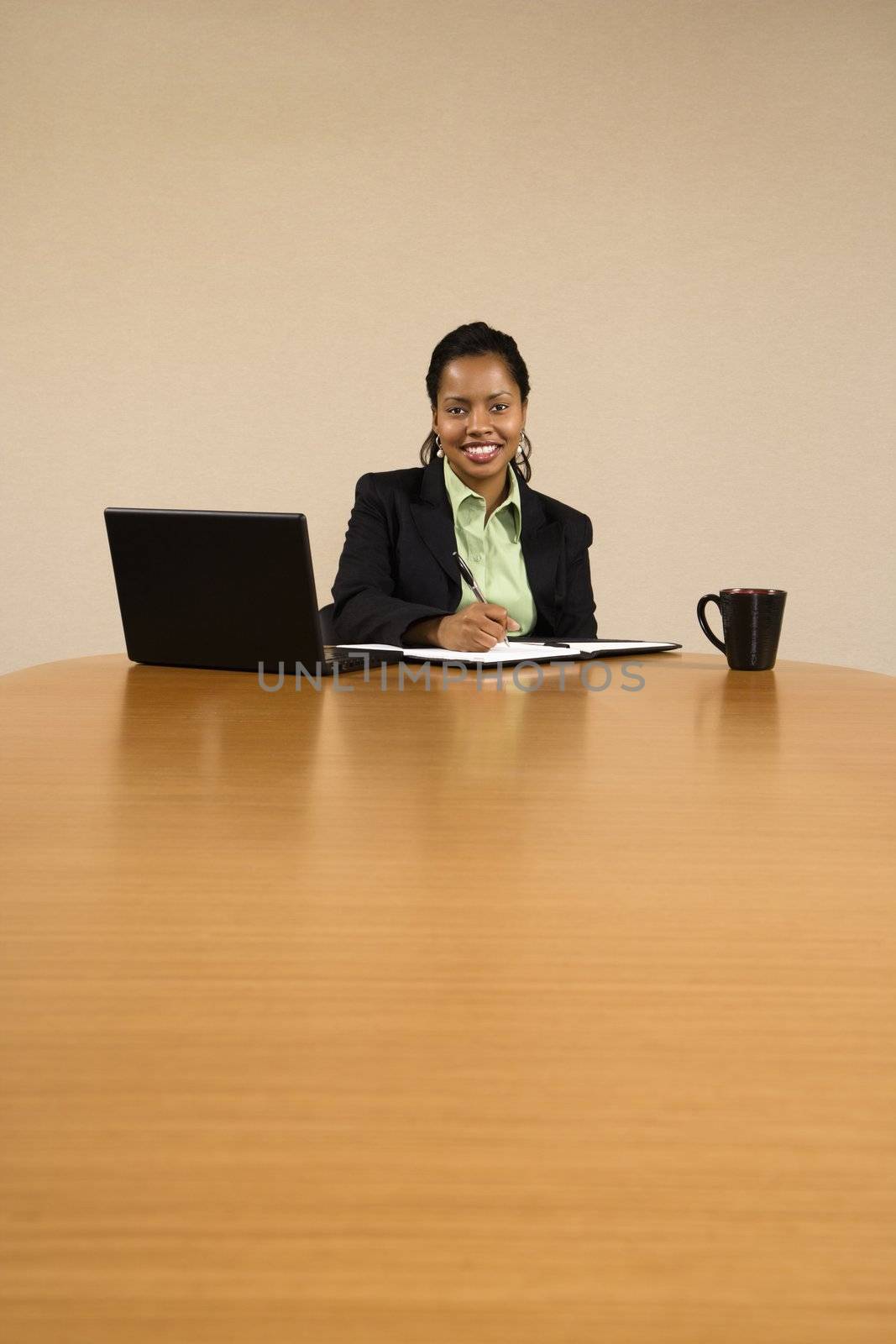 Businesswoman sitting at conference table with laptop computer and coffee cup smiling and writing in book.