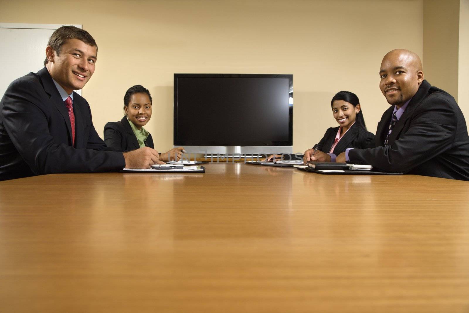 Businesspeople sitting at conference table smiling with flat screen display in background.