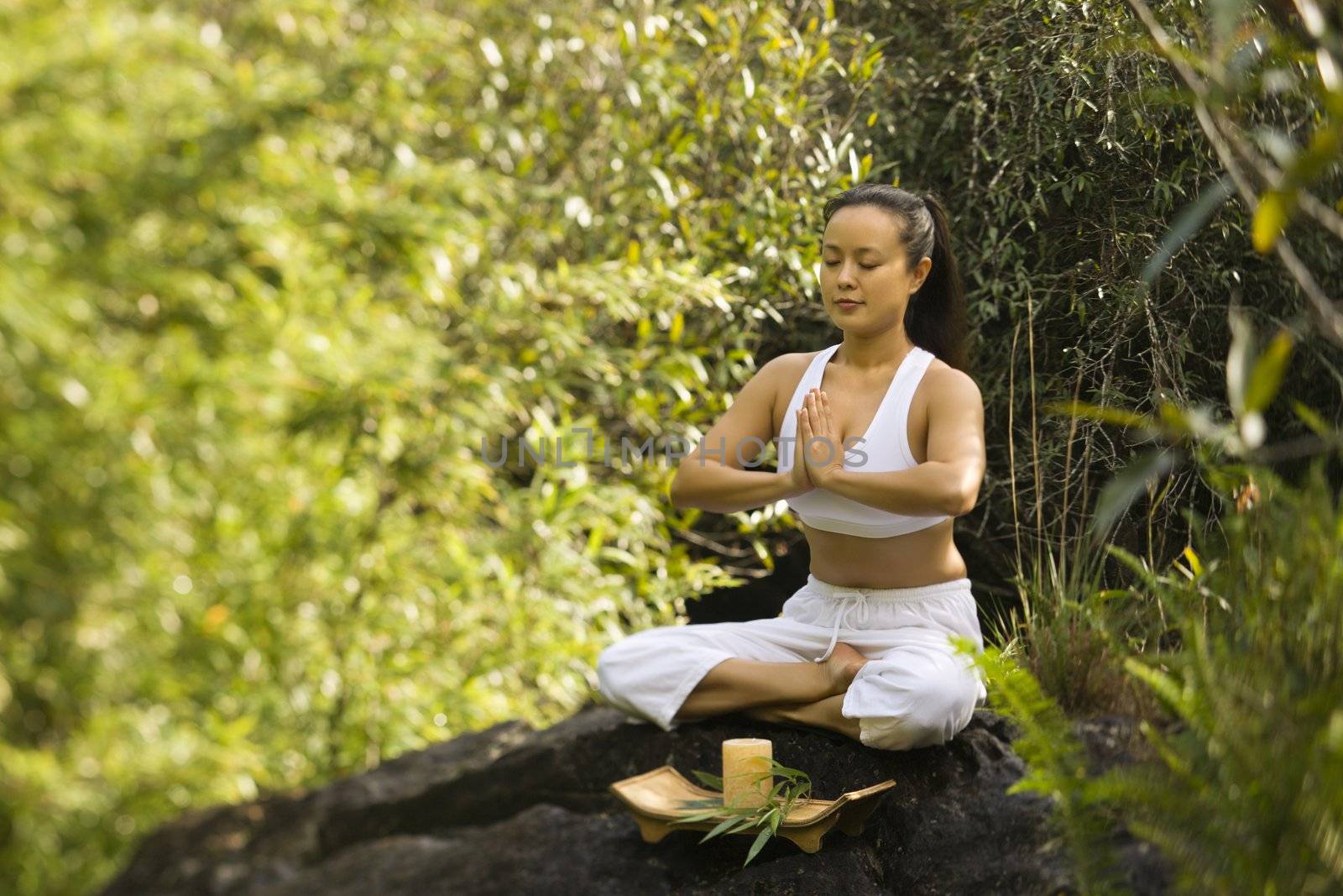 Asian American woman sitting on boulder in forest meditating with candle in Maui, Hawaii.