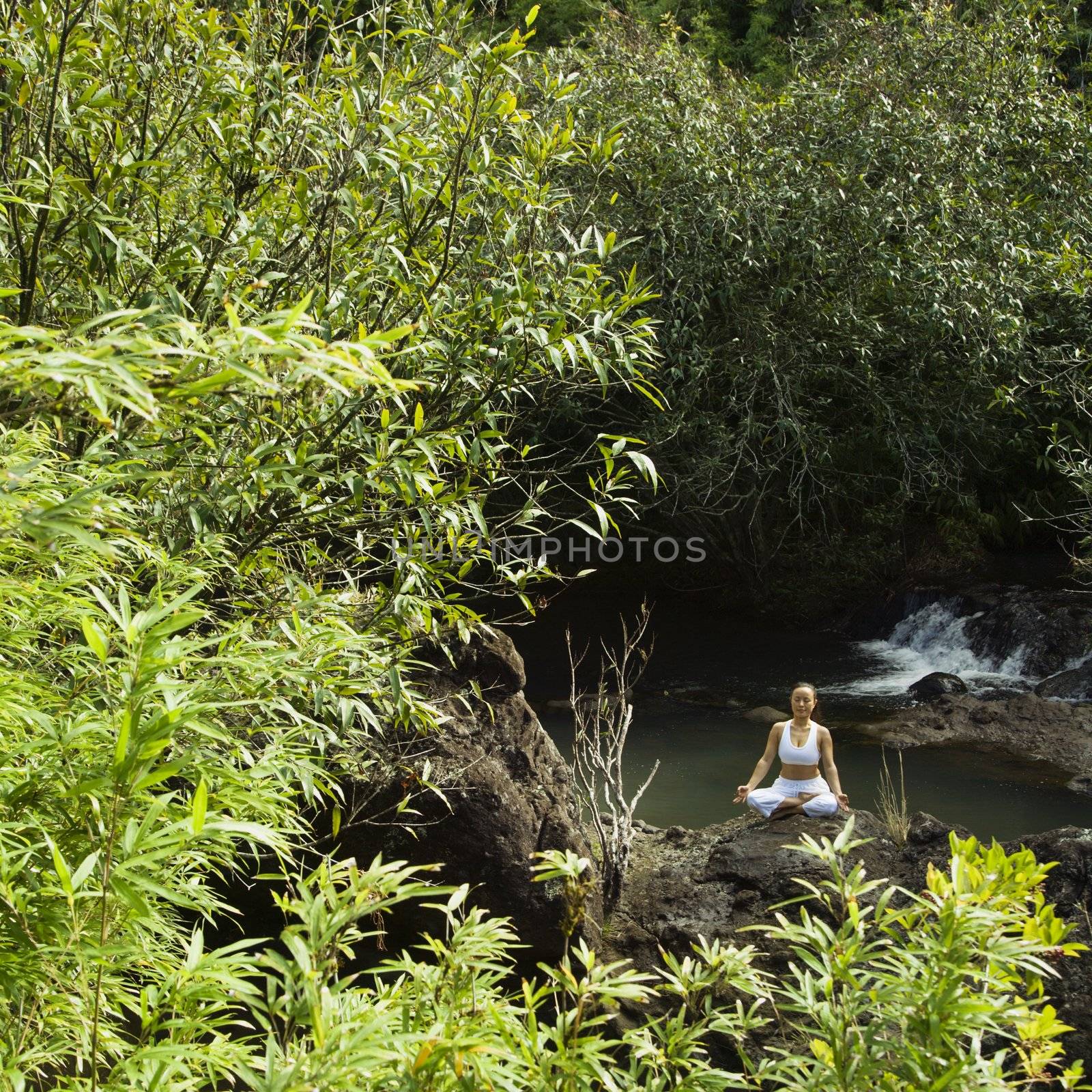 Asian American woman sitting on boulder by creek meditating in Maui, Hawaii.