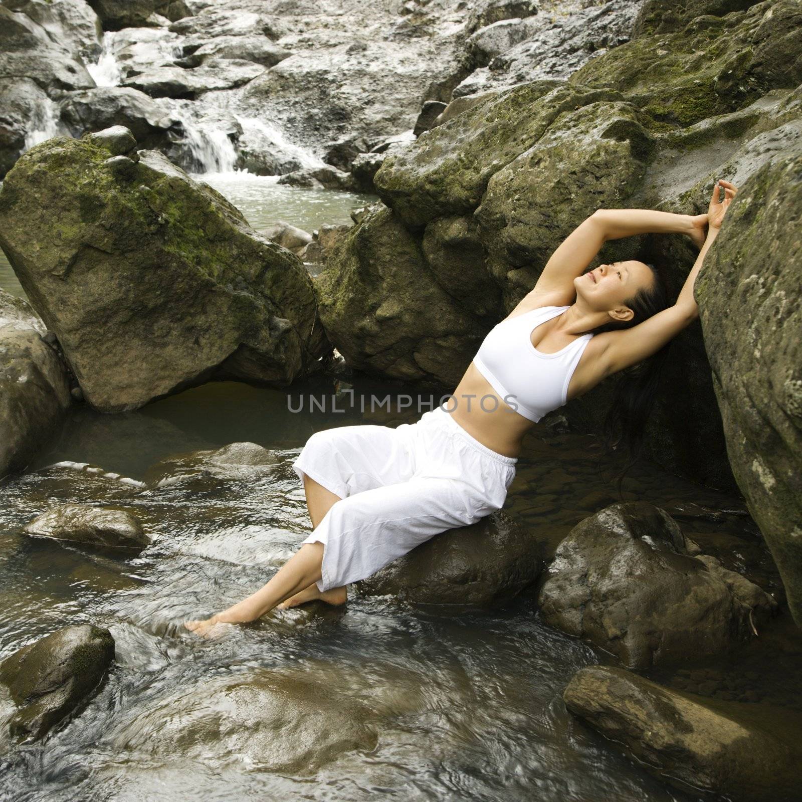 Asian American woman leaning back on boulder in creek in Maui, Hawaii.