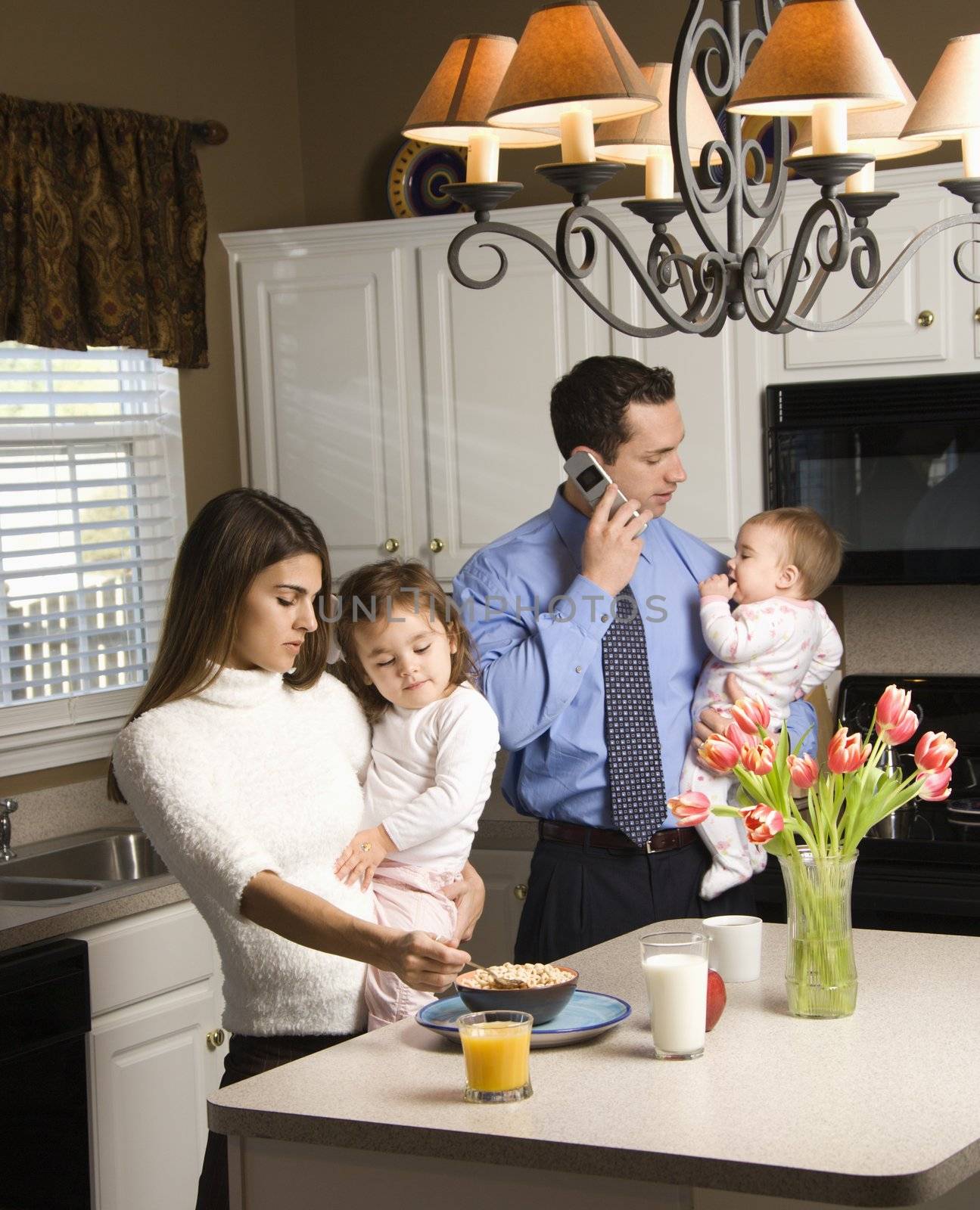 Caucasian mother and father in kitchen busy with children and cellphone.