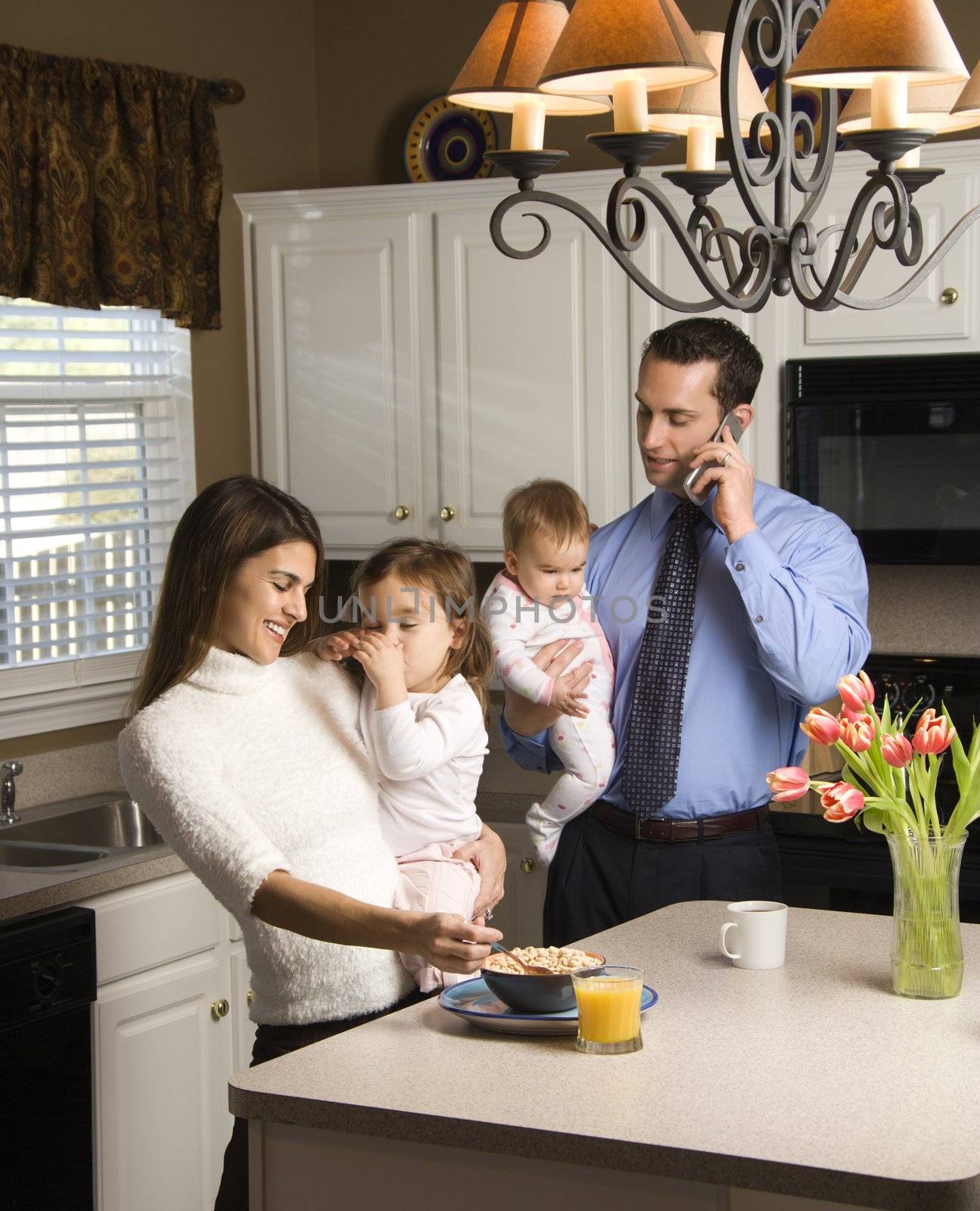 Caucasian mother and father in kitchen busy with children and cellphone.