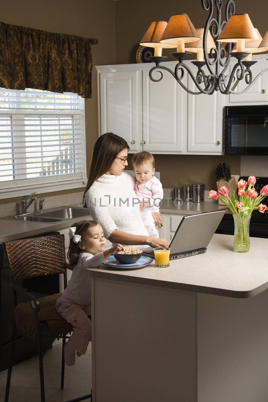 Caucasian mother holding baby and typing on laptop computer with girl eating breakfast in kitchen.