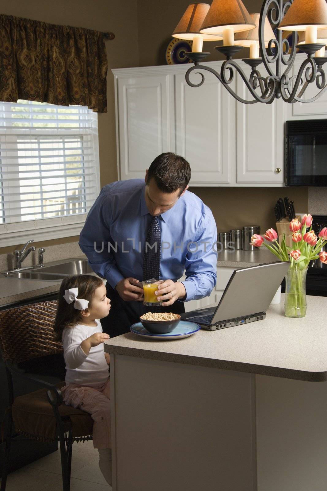 Caucasian father in suit using laptop computer with daughter eating breakfast in kitchen.