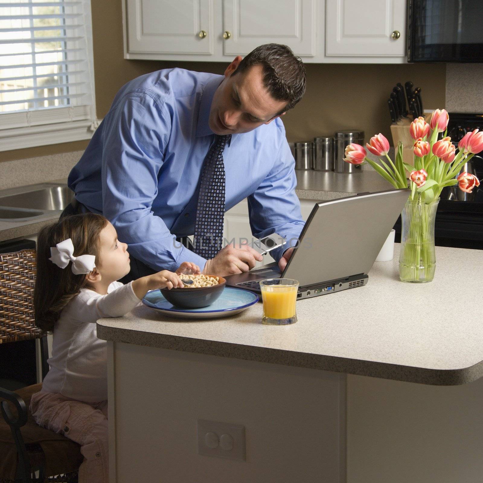 Caucasian father in suit using laptop computer with daughter eating breakfast in kitchen.
