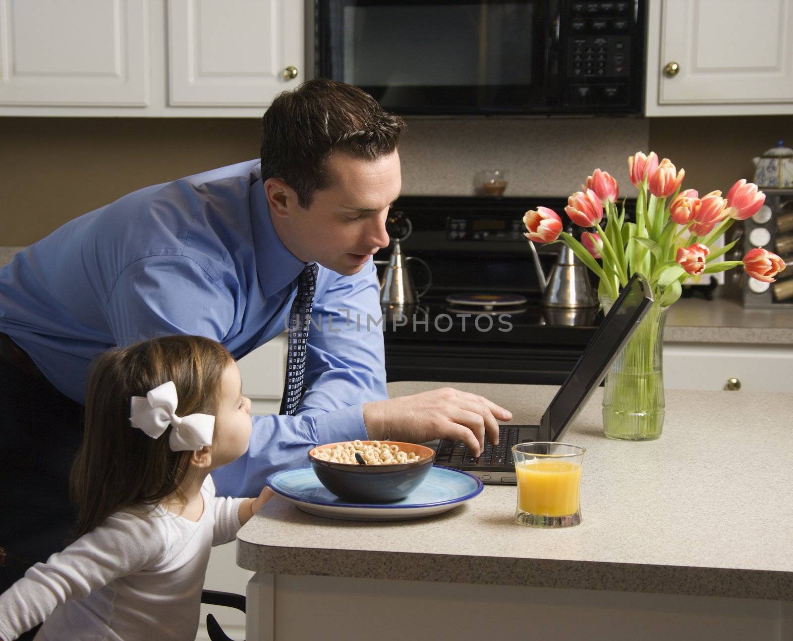 Caucasian father in suit using laptop computer with daughter eating breakfast in kitchen.