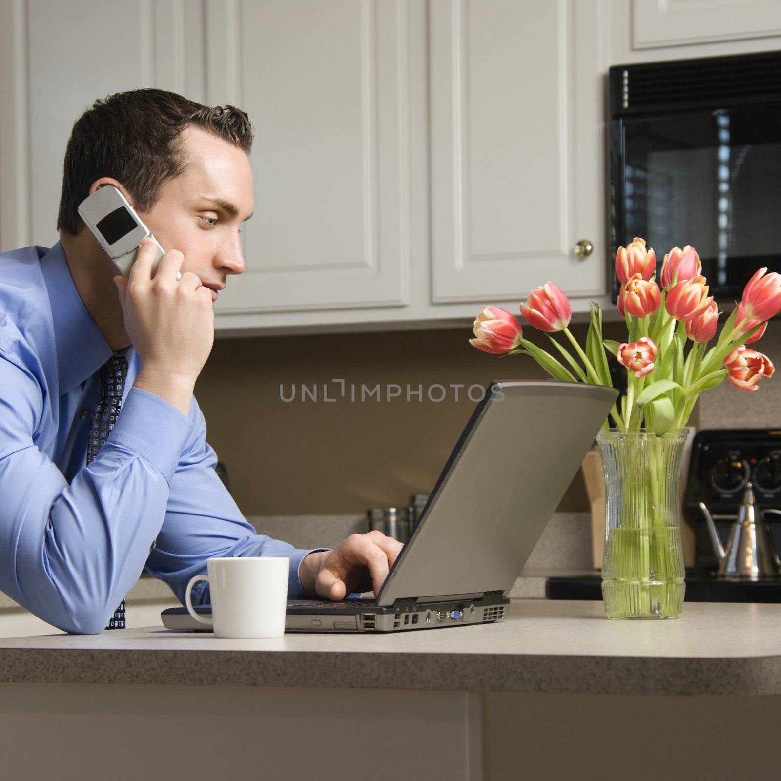 Caucasian man in suit using laptop computer and talking on cellphone in kitchen.