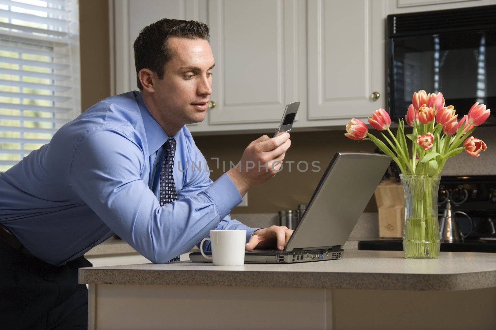 Caucasian man in suit using laptop computer and cellphone in kitchen.