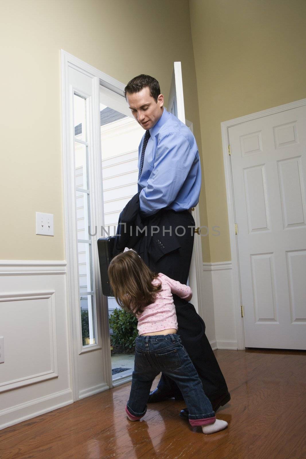 Caucasian businessman at open door with briefcase with daughter tugging on his leg.