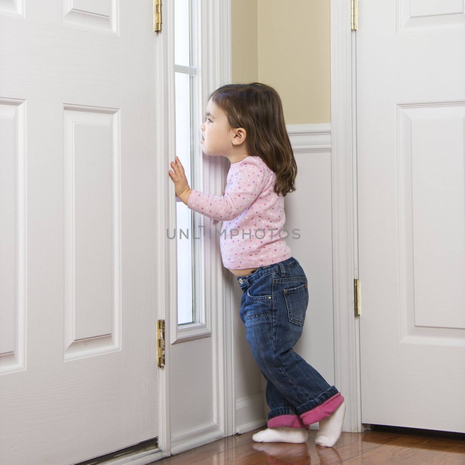 Caucasian girl toddler peeking out of window by door.