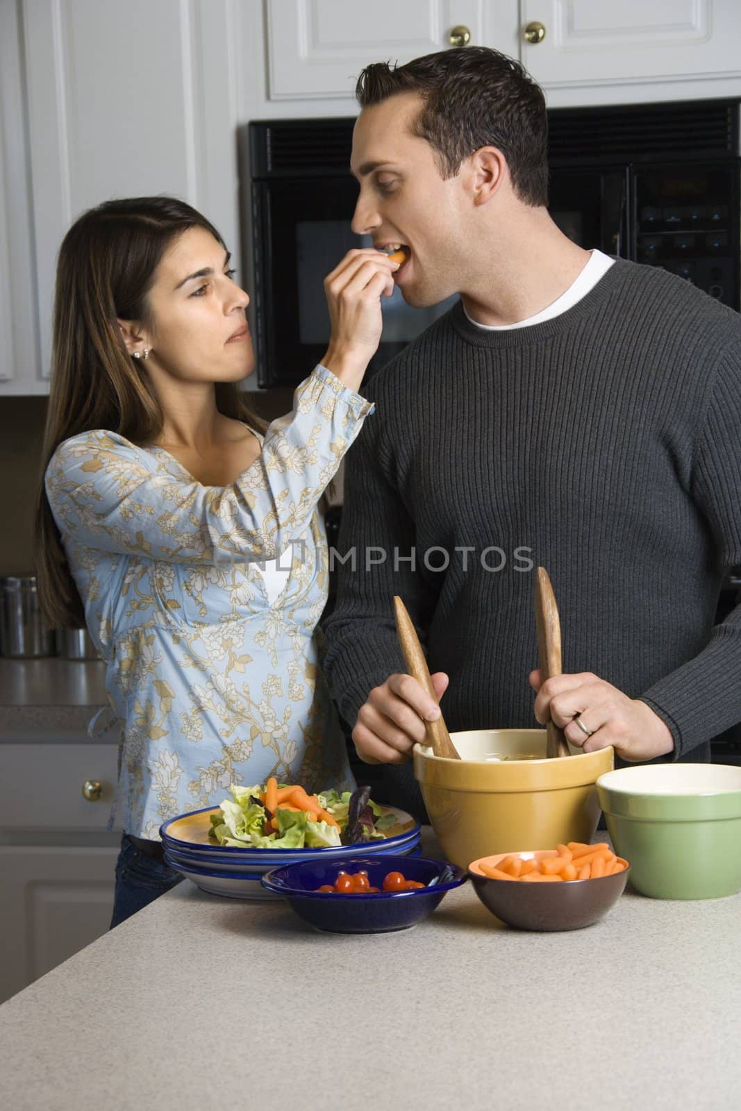 Caucasian woman feeding man at kitchen counter while he makes salad.