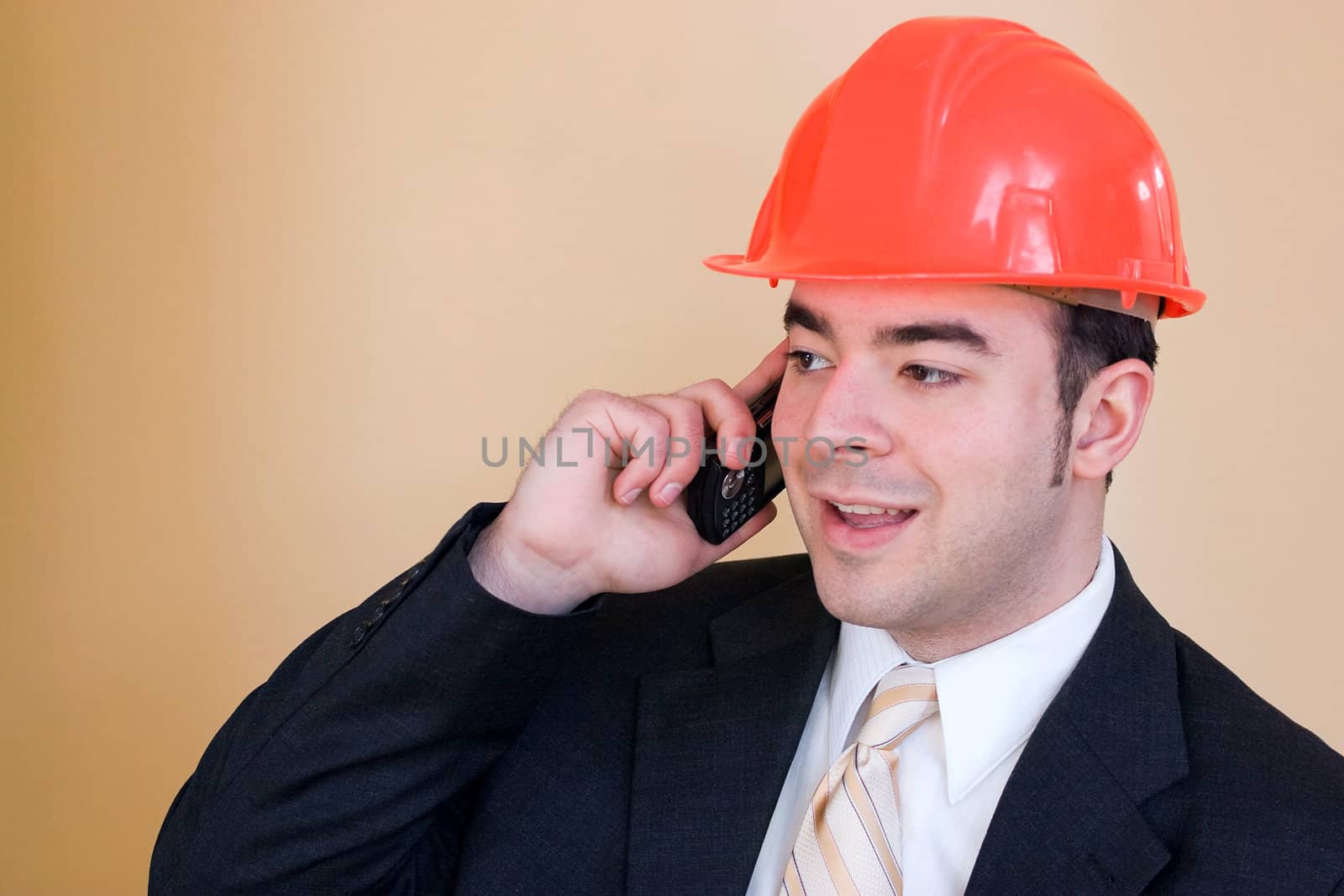 A man in a business suit and hard hat talks on his cell phone.  He could be a custom home builder or even an engineer or architect.