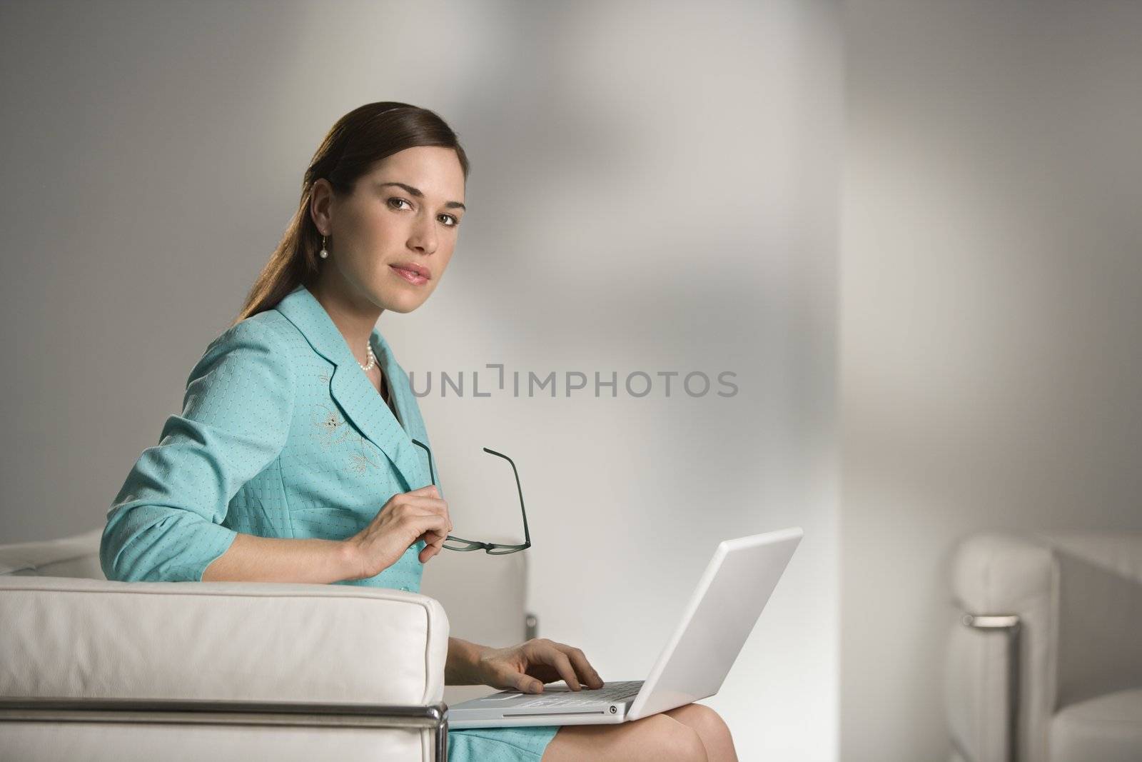 Caucasian mid adult professional business woman sitting in modern office working on laptop computer holding eyeglasses and looking at viewer.