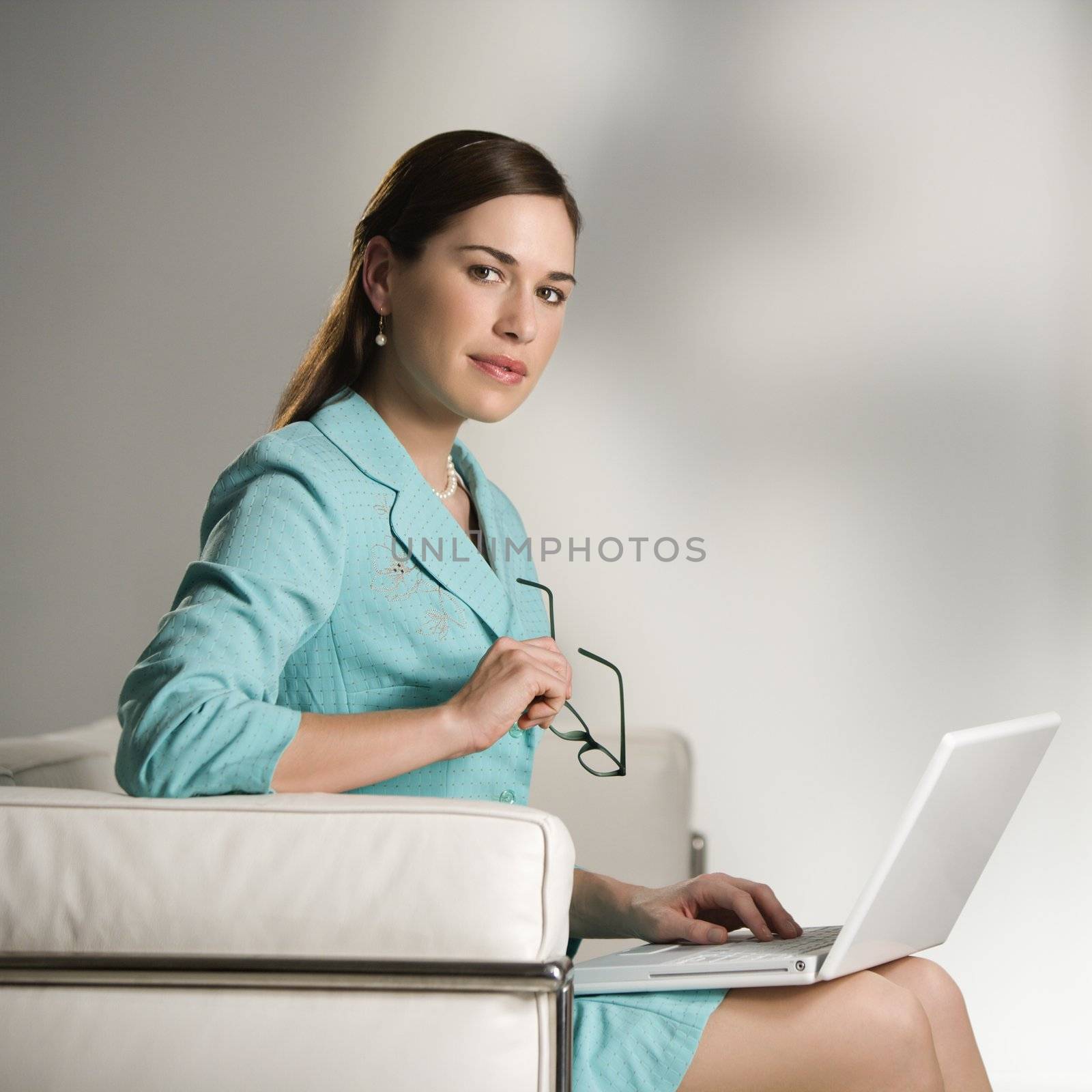 Caucasian mid adult professional business woman sitting in modern office working on laptop computer holding eyeglasses and looking at viewer.
